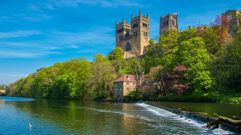 Historic stone fortress with twin towers atop a hill, overlooking a calm river and green trees along the bank.