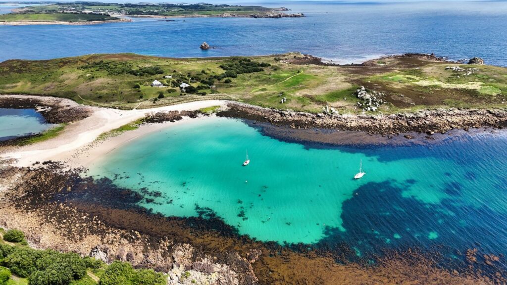 Aerial view of a green coastal landscape with a turquoise bay, sandy beach, and two sailboats on clear blue water.