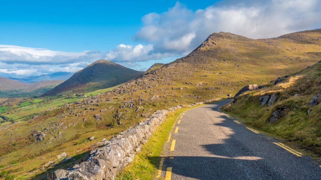 Winding rural road with a stone wall on the left, rolling hills under a blue sky in a mountainous landscape