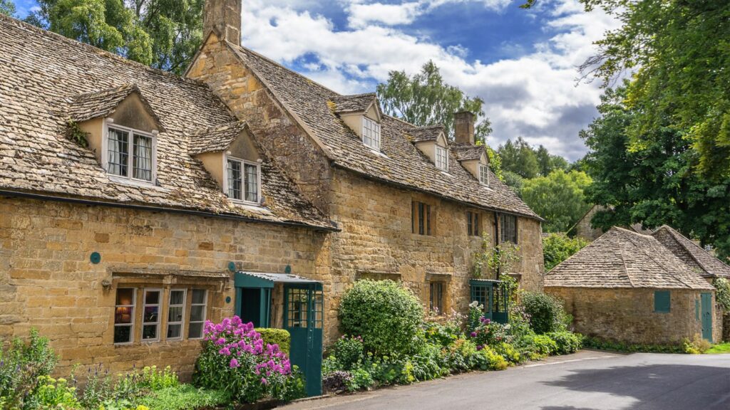 Stone village houses with steep slate roofs and blue-green doors, bordered by a colorful flower garden along a sunny road in England.