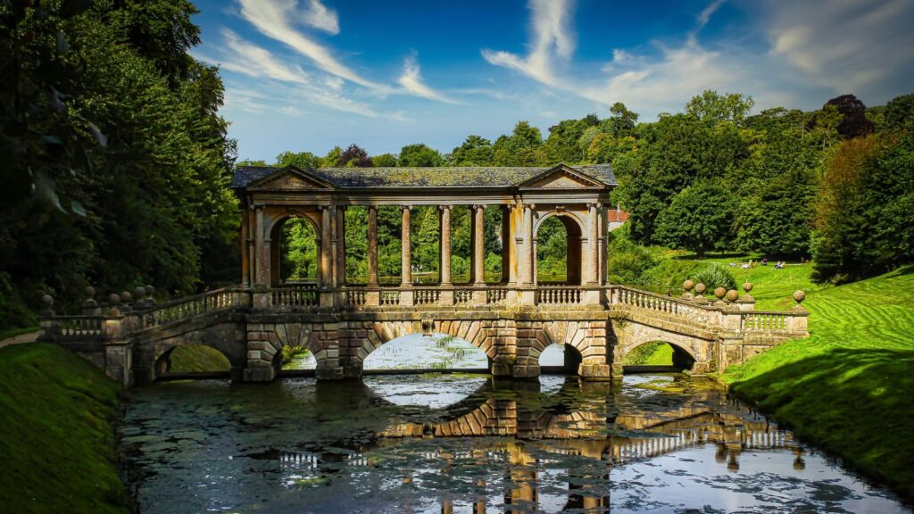 Stone, colonnaded bridge across a calm canal, surrounded by green lawns and trees under a bright blue sky.