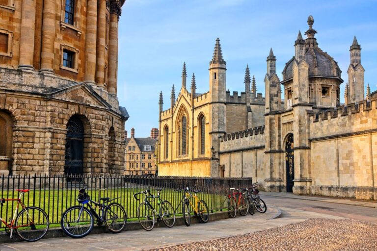 Sunlit historic stone buildings with Gothic arches and a row of bicycles parked along a black iron fence by a grassy courtyard.