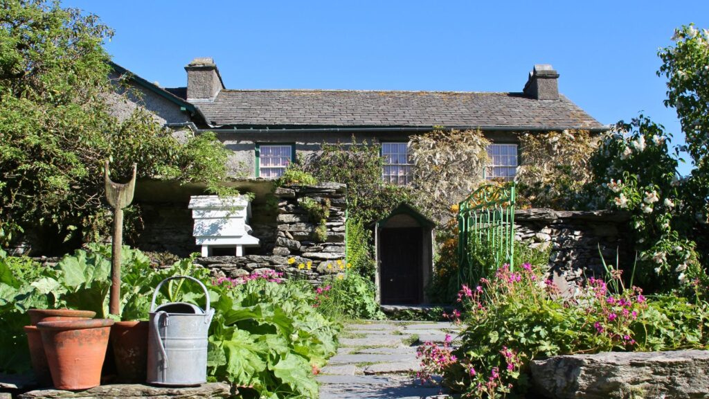 Quaint stone cottage with ivy on the walls, a stone path, and a garden filled with pots, a watering can, and a white beehive in front.