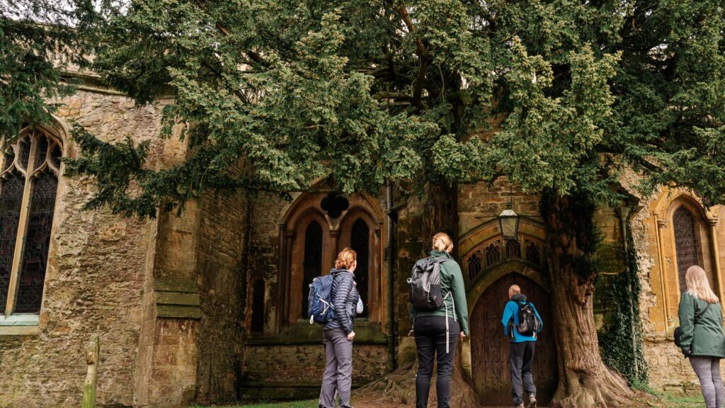 Group of hikers with backpacks approaching a historic stone church doorway beneath a large tree.